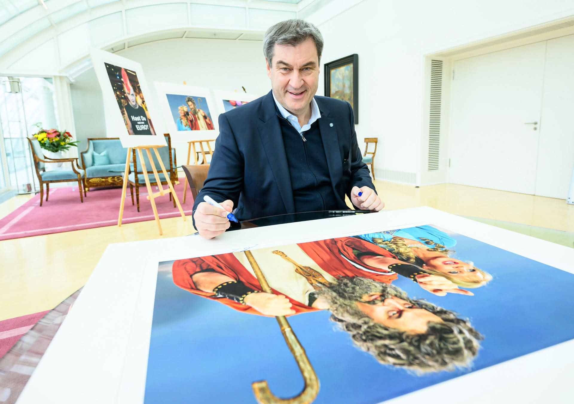 Bayerns Ministerpräsident Dr. Markus Söder signiert einen großen Kunstfotodruck auf einem Tisch, der später bei einer Charity Auktion angeboten wird, mit weiteren ausgestellten Bildern im Hintergrund.