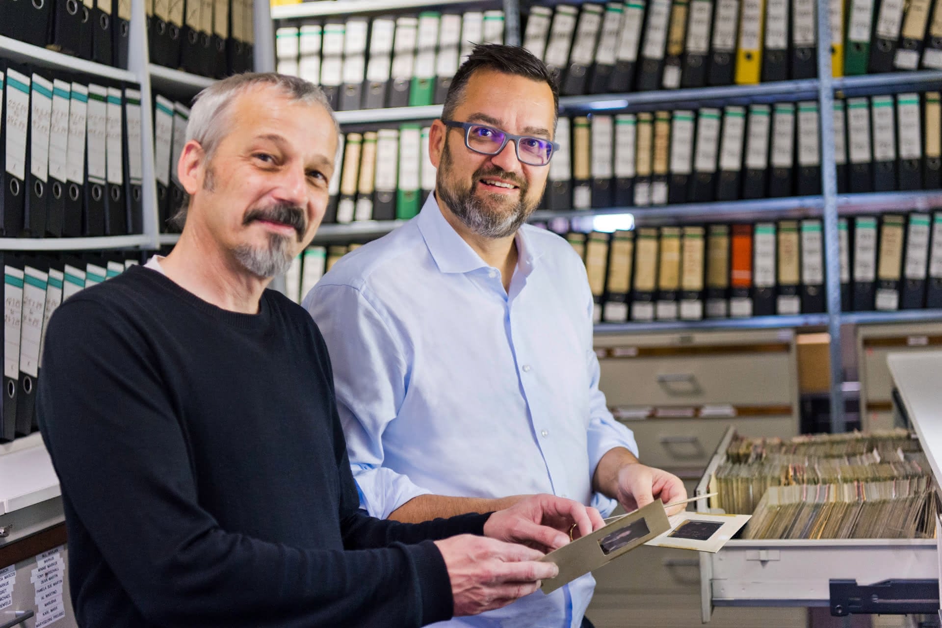 Andreas Genz, Geschäftsführer der dpa Picture-Alliance, und Christoph Grill, Leiter Content und Archivexperte, stehen zwischen hohen Regalen voller Archivmappen und betrachten historische Fotos aus den 75 Jahren der Bundesrepublik Deutschland.