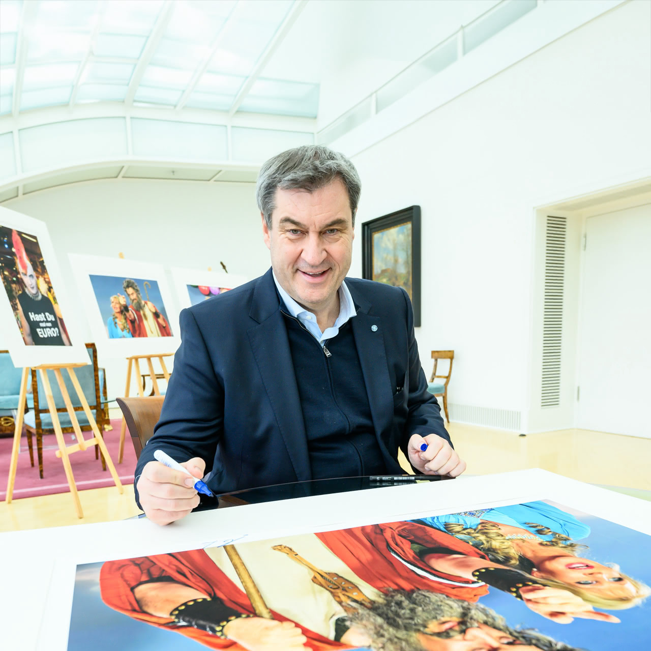 Bayerns Ministerpräsident Dr. Markus Söder signiert erste Exemplare der Fotoedition für bevorstehende Charity Auktion. Bayerns Ministerpräsident Dr. Markus Söder signiert einen großen Kunstfotodruck auf einem Tisch, der später bei einer Charity Auktion angeboten wird, mit weiteren ausgestellten Bildern im Hintergrund.
